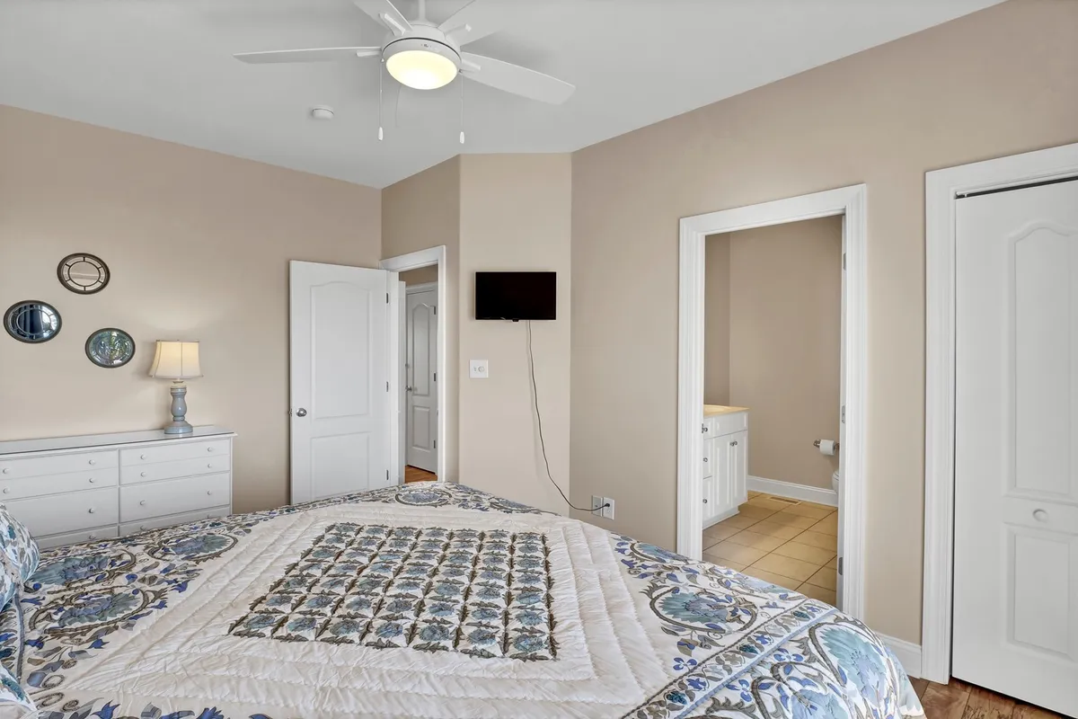This image shows a cozy guest bedroom featuring a king-sized bed adorned with a detailed quilt, a nightstand with a lamp, a wall-mounted TV for entertainment, and doorways leading to other rooms. Soft lighting and neutral tones create a comfortable, relaxing atmosphere ideal for adult guests or couples.
