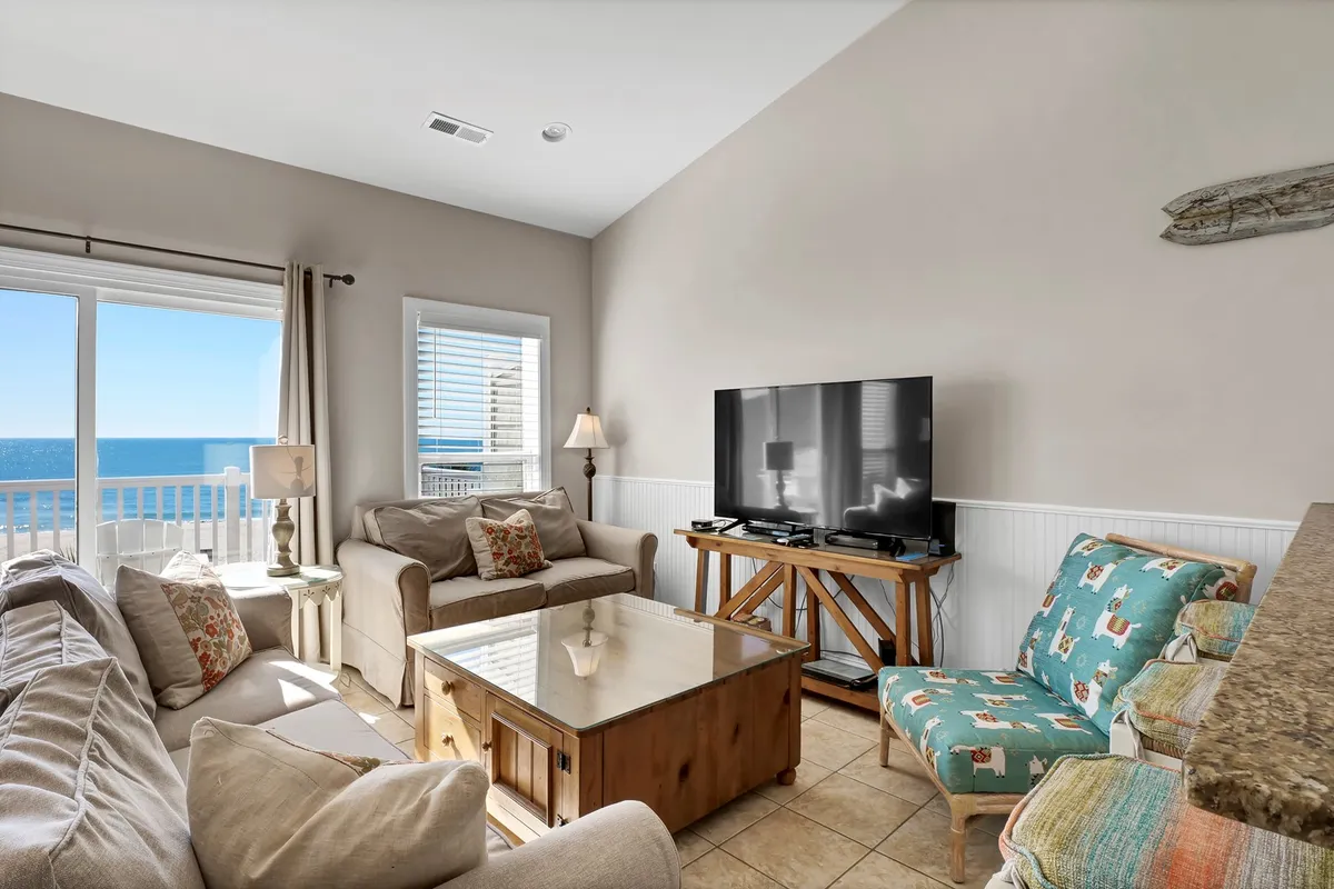 A bright and welcoming living area featuring large windows with sweeping ocean views. The space is arranged with comfortable seating, a central coffee table, and a TV, perfect for relaxing, watching movies, or gathering for conversation. Natural light fills the room, highlighting the open layout and the inviting, cheerful mood.