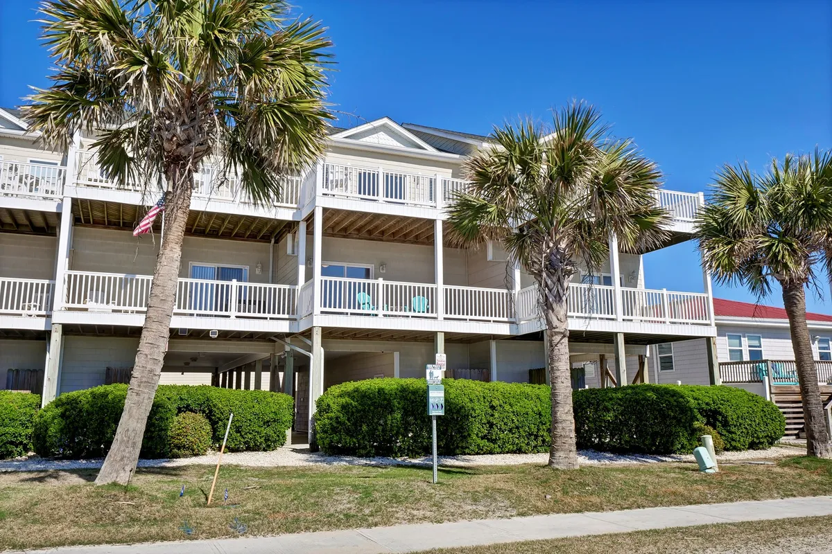 A cheerful exterior view featuring palm trees, several balconies, and lush green shrubbery. Groups of people are relaxing on balconies and enjoying the outdoors, which conveys accessibility to nature and communal outdoor living.
