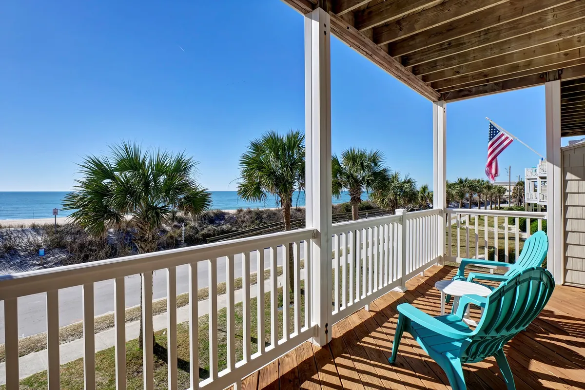 An inviting beachside deck with outdoor chairs, palm trees, and an American flag. The view stretches to the ocean, ideal for enjoying sunrises, sunsets, or watching family play on the sand below.