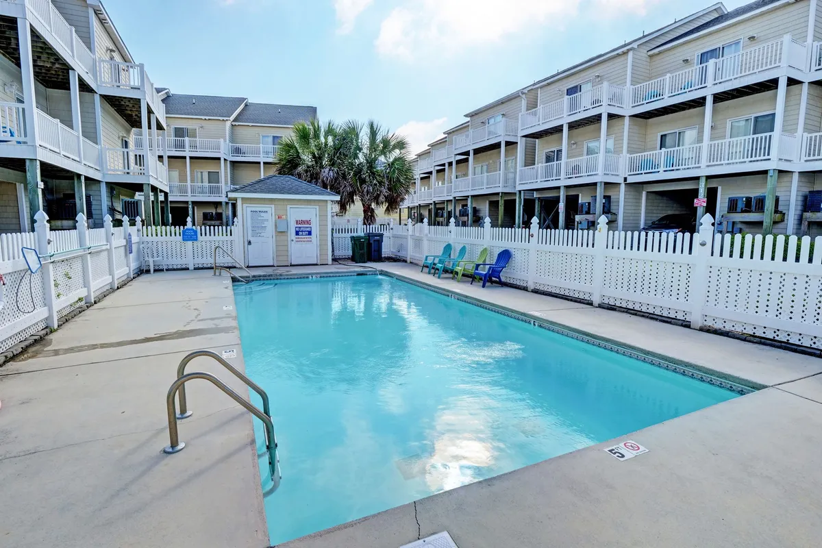 A vibrant scene of the community swimming pool surrounded by colorful lounge chairs and palm trees, with the buildings of Sea Star Village visible in the background. The pool area is sunlit, welcoming, and clearly designed for relaxation and social gatherings.