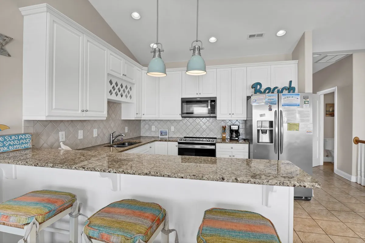 A modern kitchen with granite countertops, white cabinetry, and colorful bar stools. The space is bright and cheerful, with beach-themed decor, and is designed for both cooking and socializing, situated adjacent to the living area.
