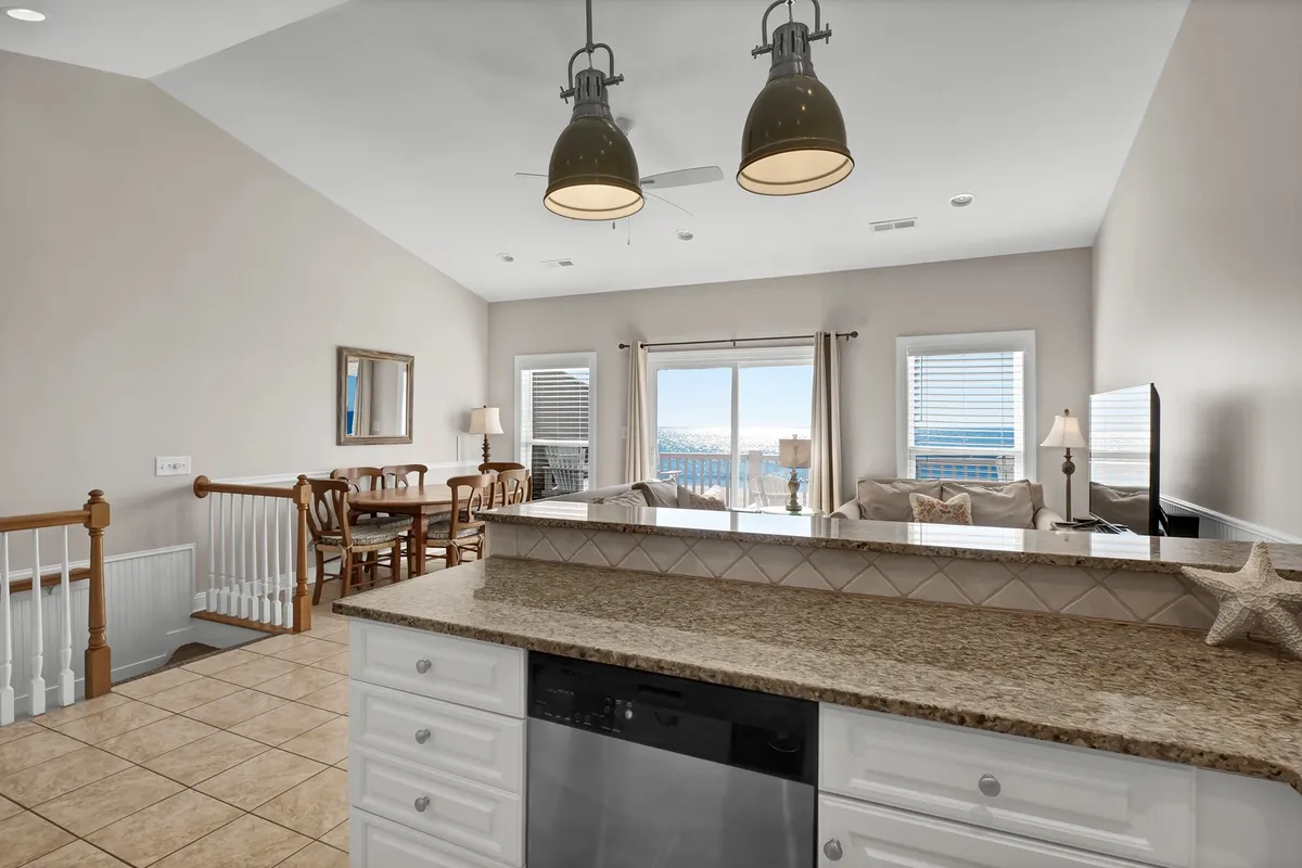 A welcoming dining area and kitchen with granite countertops, large windows, and a view of the living area. The space is bright and inviting, featuring a spot for meals, socializing, and watching sunsets through the windows.
